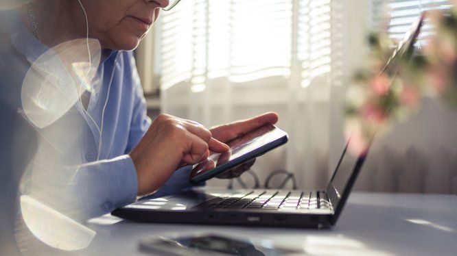 personne assise à un bureau tenant un smartphone dans le main devant un écran d'ordinateur portable