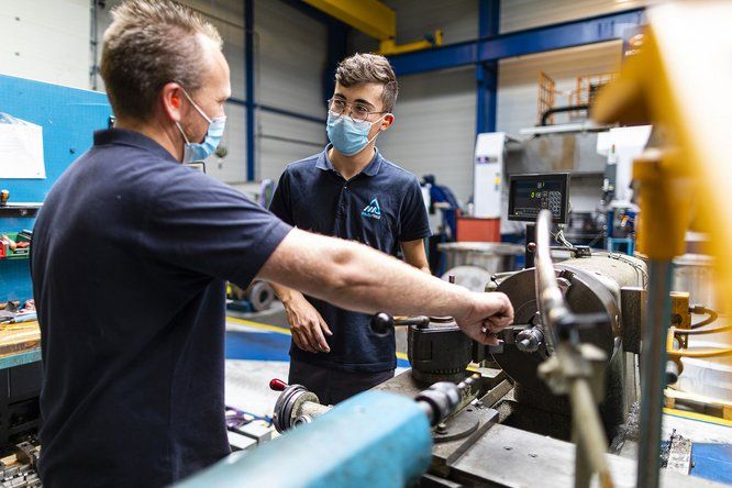 Un homme masqué discute avec son jeune apprenti masqué à côté de machines industrielles