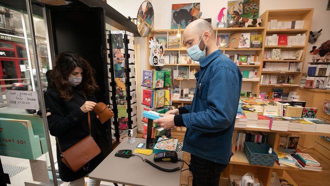 Une cliente masquée dans une librairie face au libraire masqué, un comptoir rempli de livres les séparent.