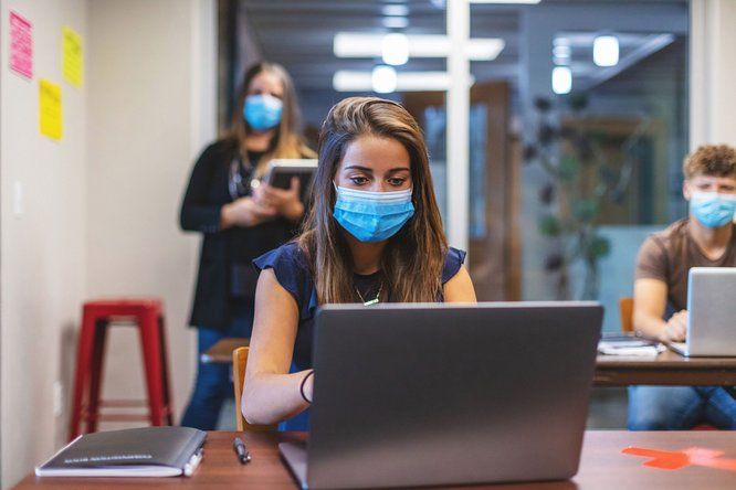 Une jeune lycéenne portant un masque fixe l'écran de son ordinateur posé sur un bureau en bois